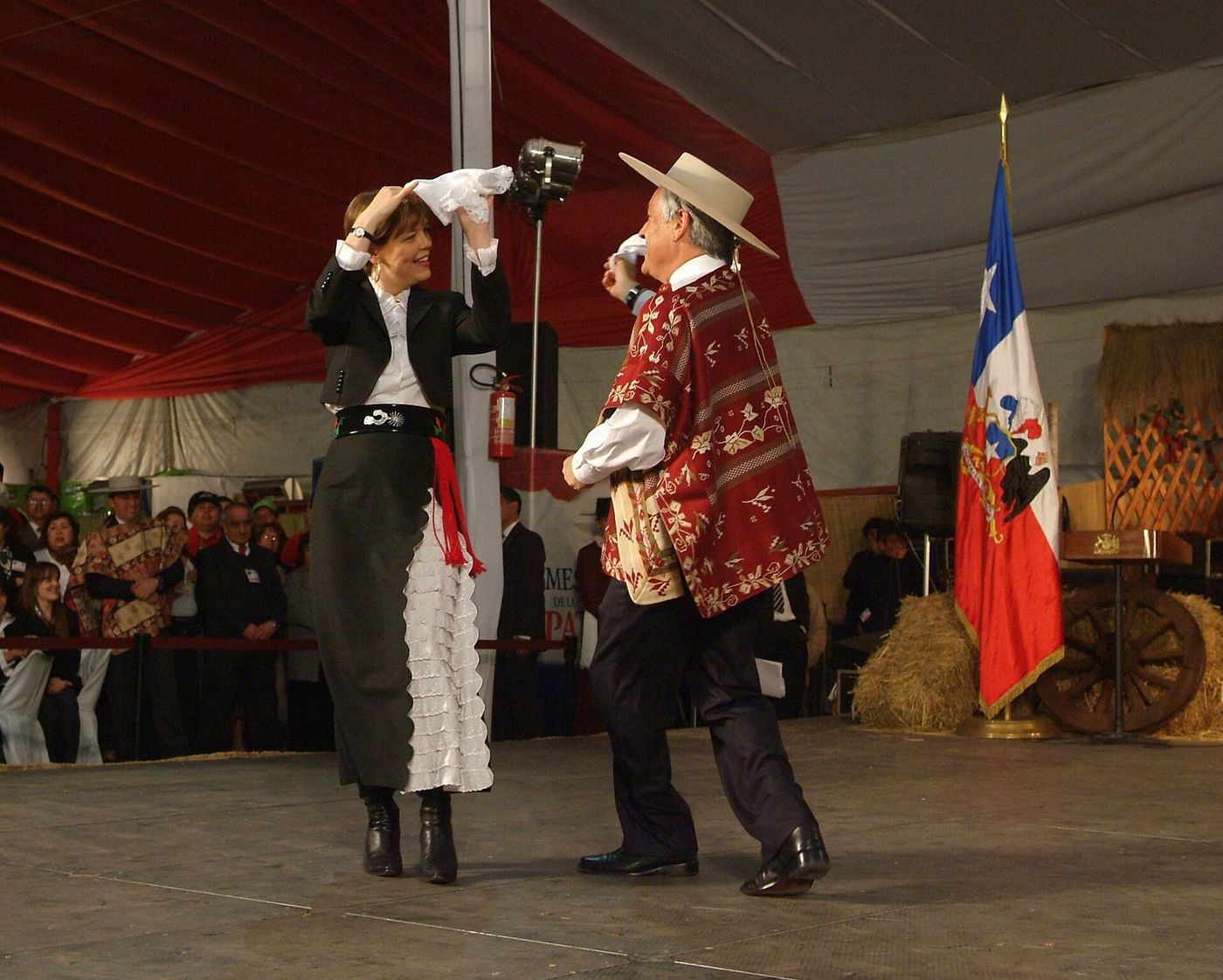A couple dancing cueca with handkerchiefs inside a fonda beside a Chilean flag