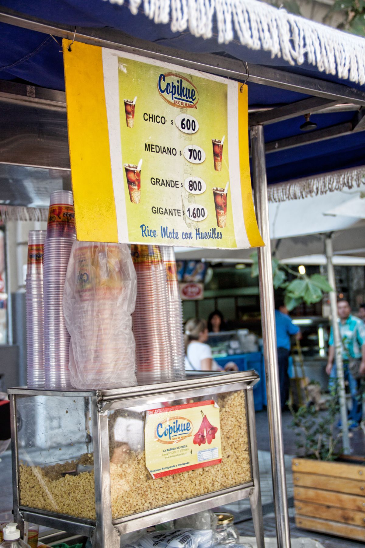 A street cart selling mote con huesillo with a price board listing cup sizes