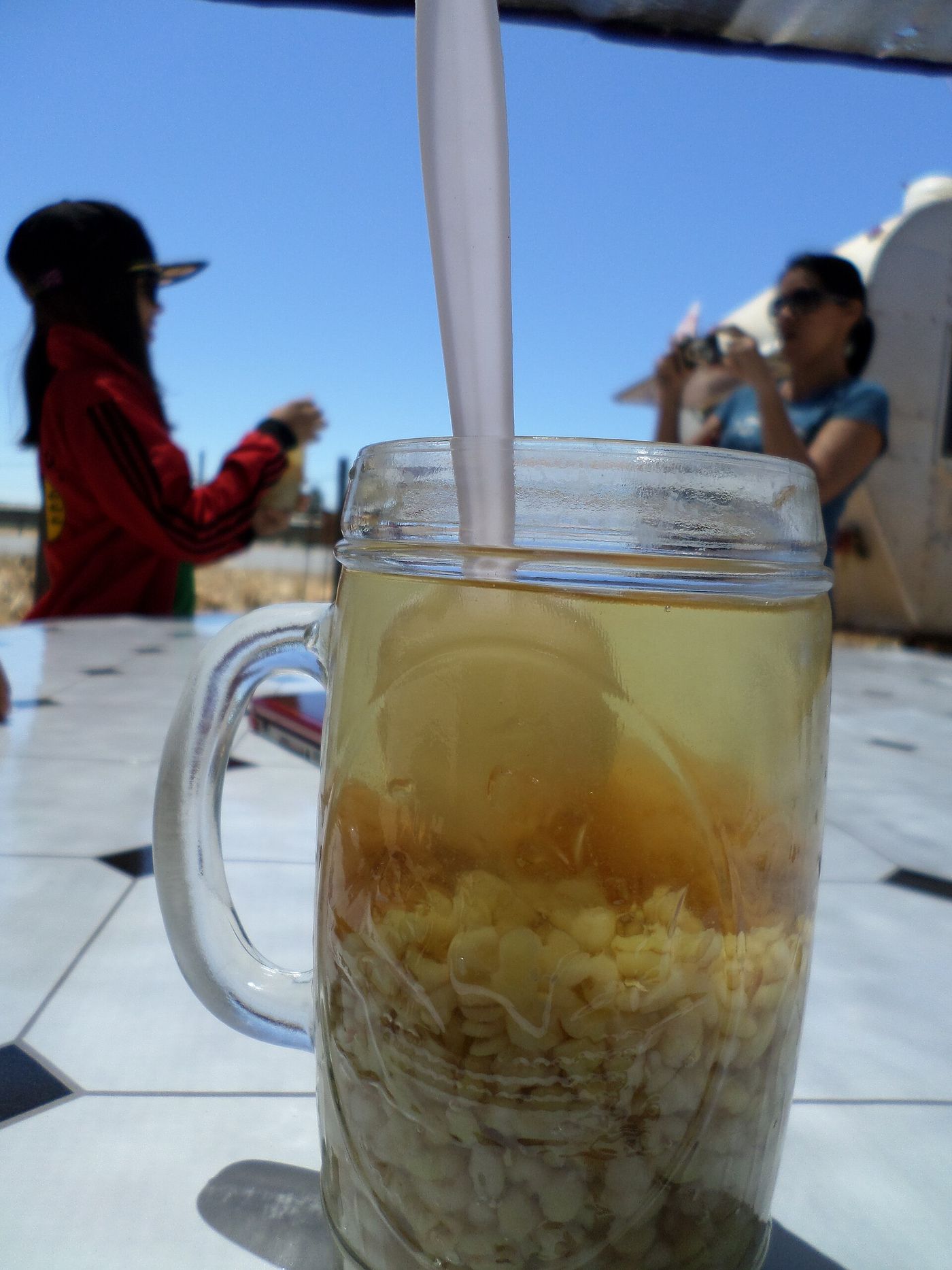 A glass of mote con huesillo with husked wheat and a dried peach in syrup, held outdoors in the sun