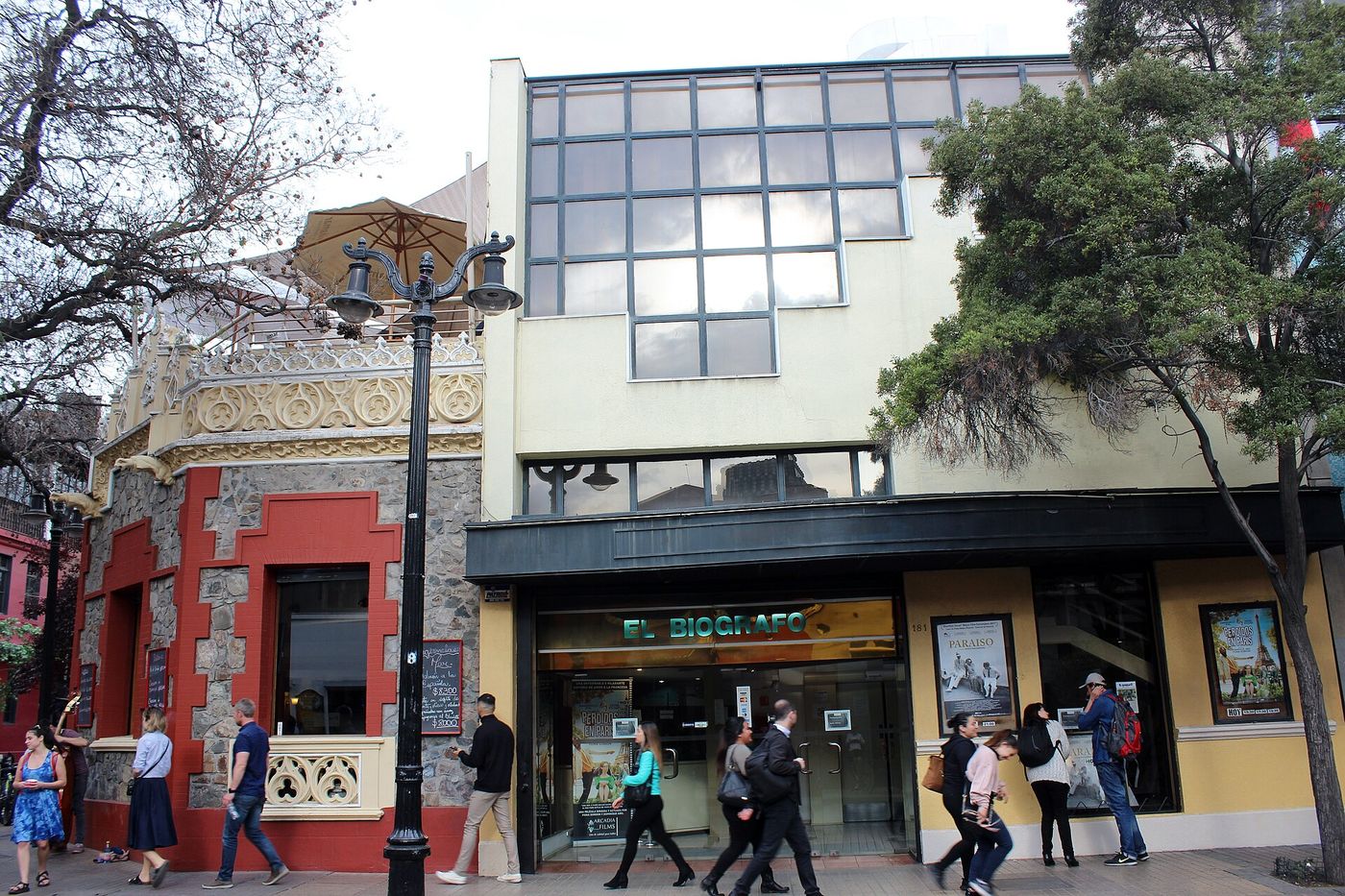 Pedestrians in jackets passing the El Biografo cinema on Calle Lastarria under bare winter trees
