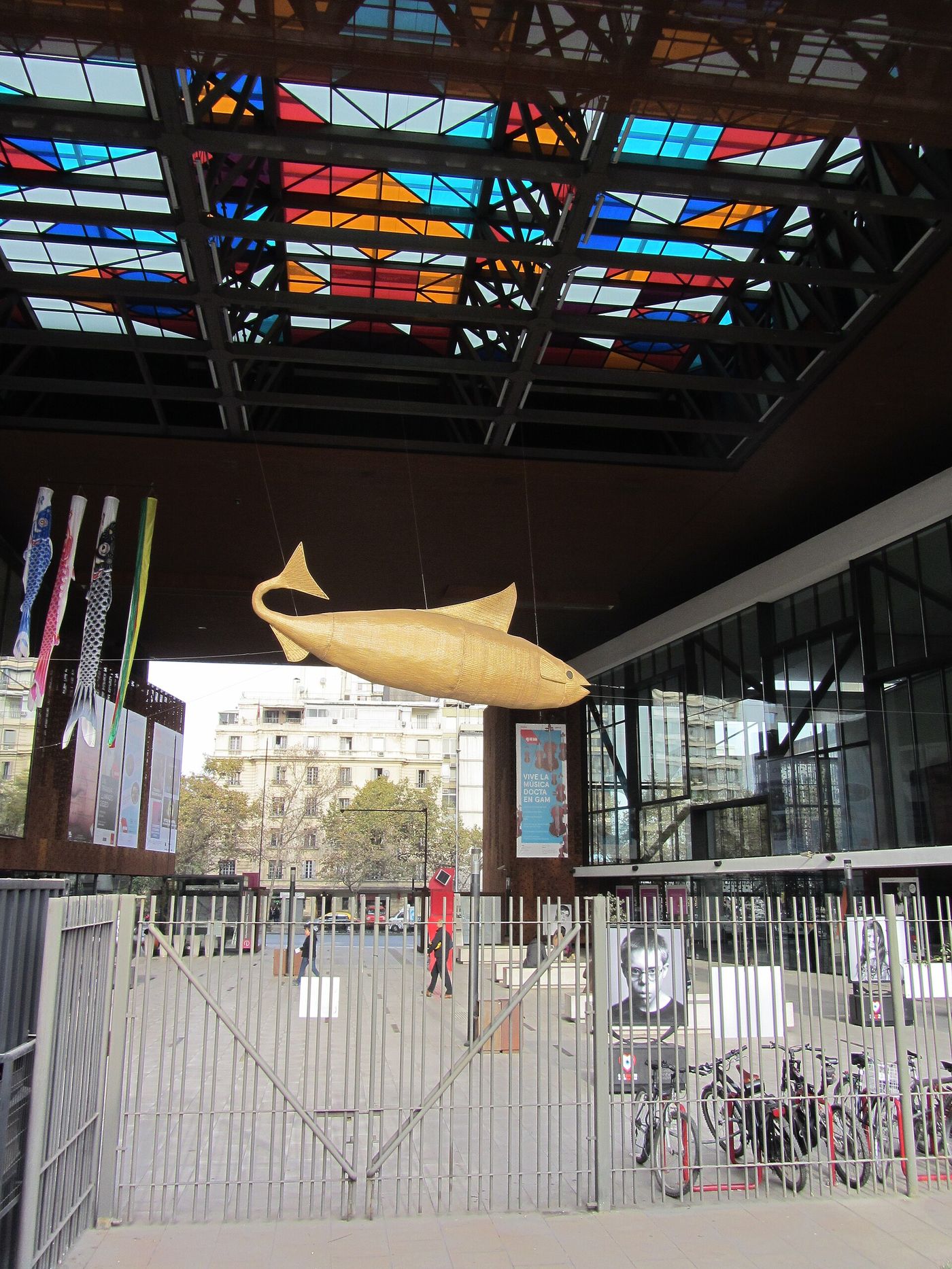 A wicker fish sculpture hanging under the coloured glass canopy of the GAM plaza in Santiago