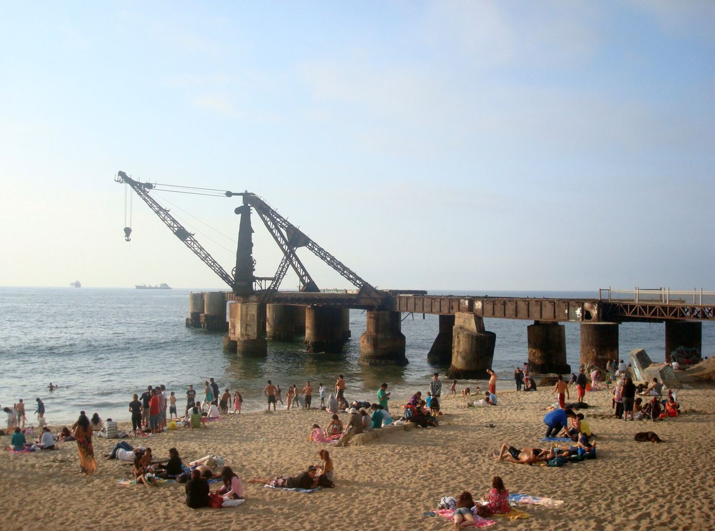 Beachgoers on the sand in front of the old iron Muelle Vergara pier with its crane, on a sunny afternoon