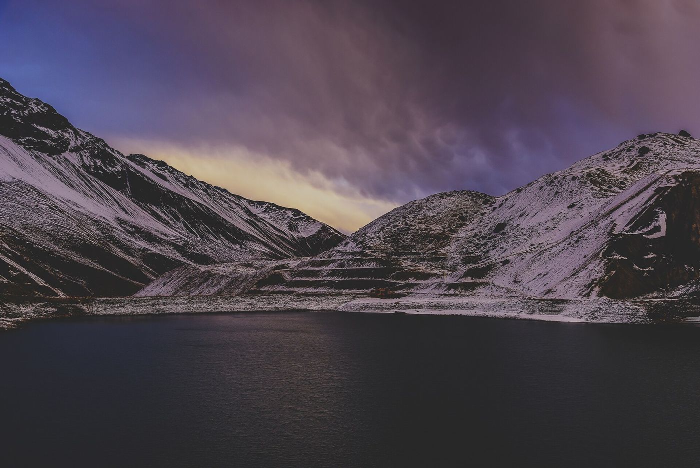 Dark water of Embalse El Yeso under storm clouds, with snow on the surrounding slopes