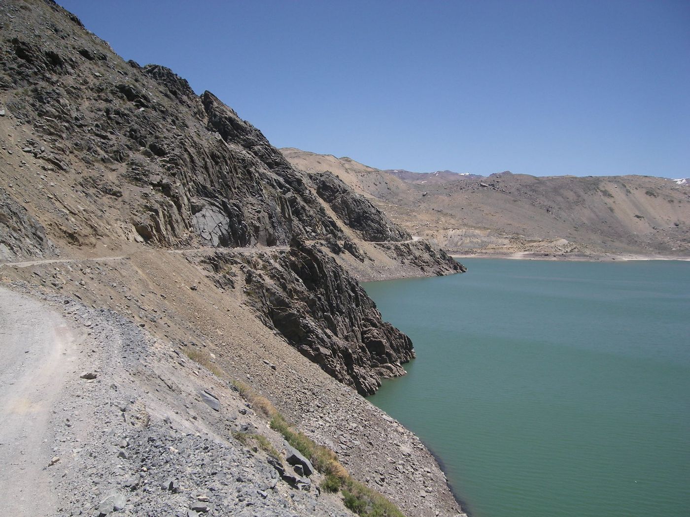 A gravel road cut into a bare slope above the green-blue water of Embalse El Yeso