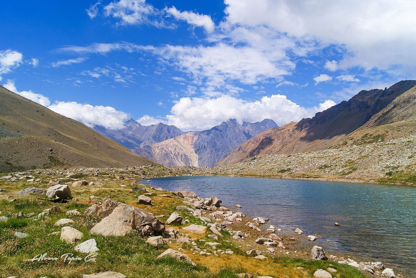 The El Morado lagoon below colorful mountains on a sunny day, with rocks and grass on its shore