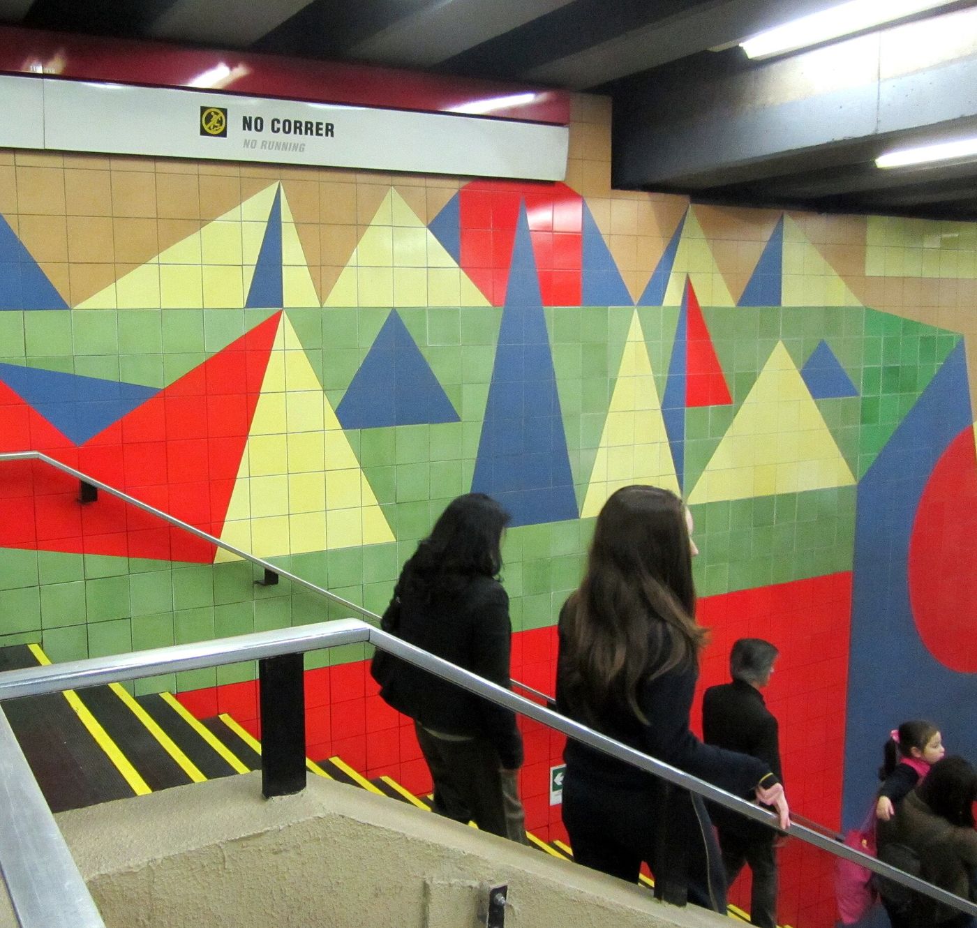 Passengers walking down stairs past a colorful geometric tile mural in Los Leones Metro station