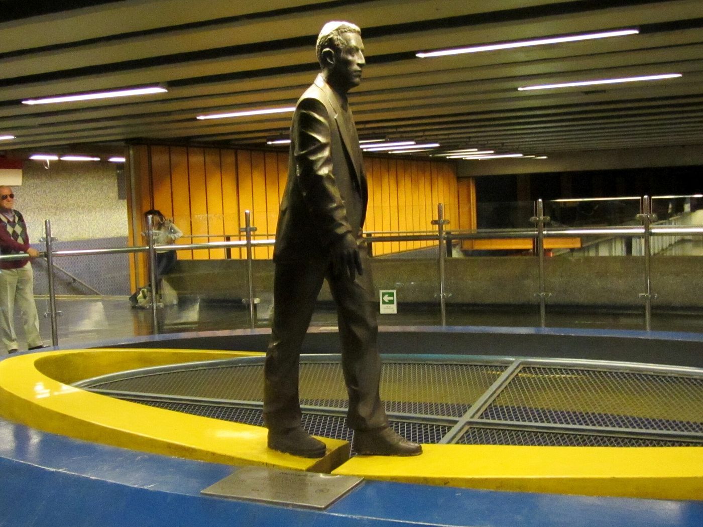 A bronze statue of a walking man on a yellow and blue platform inside Metro Universidad Católica station