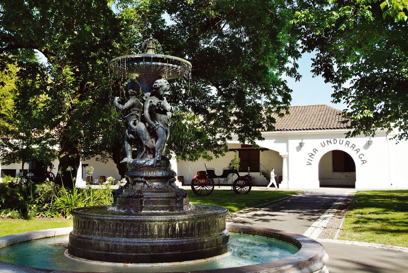 An ornate iron fountain in front of the whitewashed entrance arch of Viña Undurraga, with a horse-drawn carriage beyond